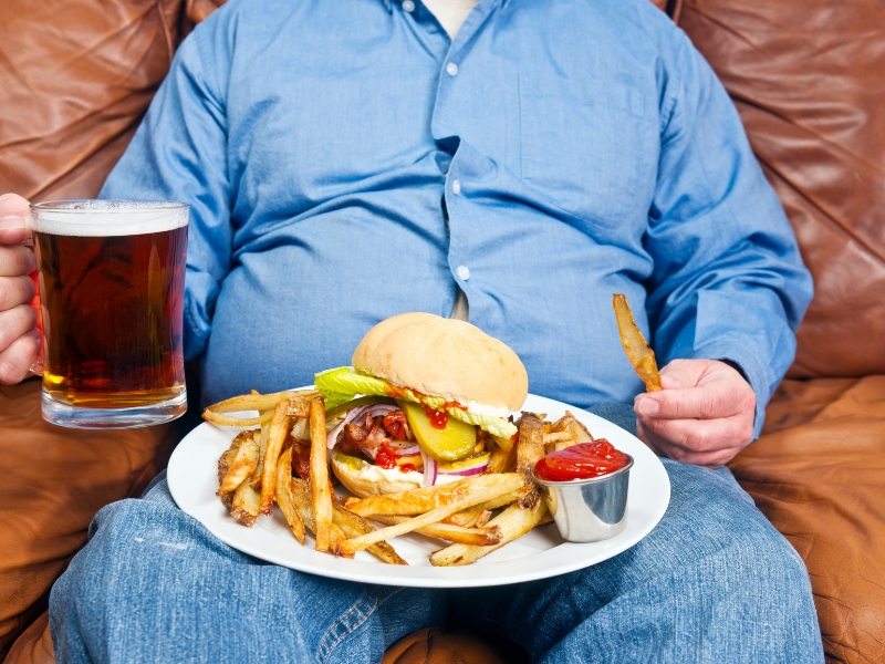 Overweight person eating a burger, fries, and beer — examples of the worst foods for slow metabolism.