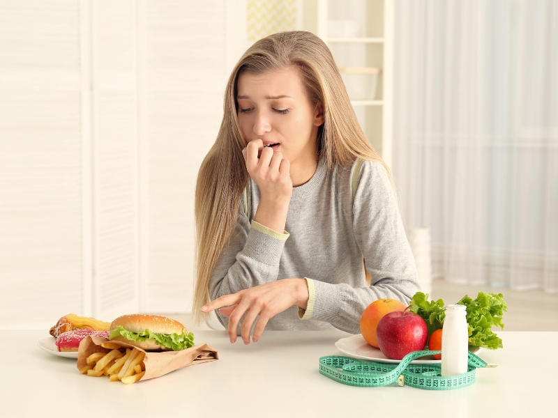 Young woman hesitating between cheat meals like burgers and fries or healthy options like apples, lettuce, and yogurt.