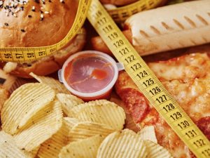 Unhealthy cheat meals, including burgers, pizza, chips, and sauce, with a measuring tape symbolising diet struggles.