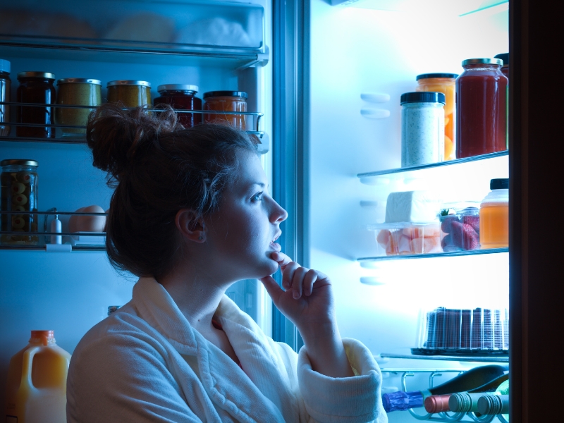 A woman in a bathrobe ponders late-night cravings while looking into a fridge filled with juices, dairy, and desserts.